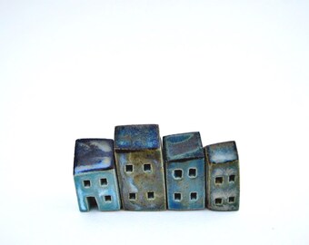 Puede incluir: Un conjunto de cuatro pequeñas figuras de casas de cerámica dispuestas en fila. Cada pieza presenta un esmalte brillante y moteado en tonos azul profundo, cerceta y marrón tierra, con pequeñas aberturas cuadradas para las ventanas y una puerta.