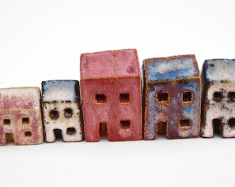 Puede incluir: Una fila de cinco pequeñas figuras de casas de cerámica con superficies texturizadas. Las casas presentan esmaltes variados en tonos rosa, rojo, azul y blanco, cada una con recortes cuadrados que representan ventanas y puertas.