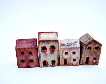 Puede incluir: Cuatro pequeñas figuras de casas de cerámica dispuestas en fila. La primera es de color rojo intenso, la segunda crema con detalles rojos, la tercera rosa claro con blanco y la cuarta es una casa de tonos morados con techo a dos aguas y ventanas.