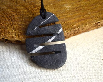 Puede incluir: Un colgante de piedra gris oscuro con un cordón negro. El colgante tiene un diseño único con cortes horizontales y vetas blancas. El fondo es una superficie de madera y una tela blanca.