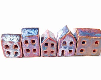 Puede incluir: Una fila de cinco pequeñas figuras de casas de cerámica con un acabado esmaltado en tonos azul, morado y marrón rojizo. Cada casa presenta ventanas cuadradas recortadas y una forma de techo distinta, dispuestas en línea.