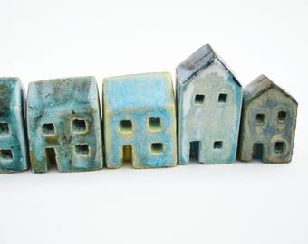 Puede incluir: Un conjunto de cinco pequeñas figuras de casas de cerámica dispuestas en fila. Cada casa presenta un acabado esmaltado único en tonos azul, verde azulado y blanco, con ventanas y puertas cuadradas recortadas en la estructura.
