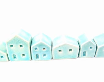 Puede incluir: Un conjunto de seis pequeñas figuras de casas de cerámica con un esmalte azul turquesa claro. Cada casa presenta sencillos recortes cuadrados que representan ventanas y puertas, con formas de techo y tamaños variados en una fila.