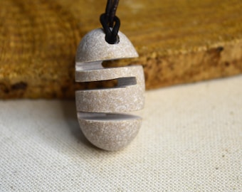 Puede incluir: Colgante de piedra beige claro con un diseño tallado en capas. El colgante está suspendido de un cordón negro. El fondo presenta una superficie de madera marrón texturizada y tela blanca.