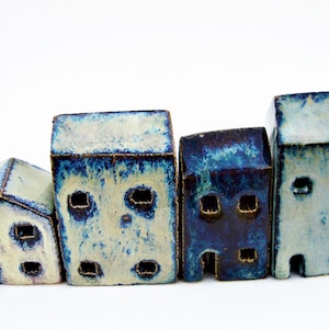 May include: A set of four ceramic house figurines with a glossy glaze in shades of blue, white, and cream. Each structure features small square cutouts for windows and a simple arched opening for a door, with varying heights and roof shapes.