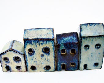 Puede incluir: Un conjunto de cuatro figuras de casas de cerámica con un esmalte brillante en tonos azul, blanco y crema. Cada estructura presenta pequeñas aberturas cuadradas para las ventanas y una sencilla apertura arqueada para la puerta.