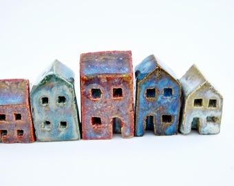 Puede incluir: Un conjunto de cinco pequeñas figuras de casas de cerámica dispuestas en fila. Cada casa presenta un acabado esmaltado único en tonos azul, rojo y beige, con ventanas y puertas cuadradas recortadas. Las estructuras varían en altura y anchura, mostrando un aspecto texturizado y artesanal con superficies brillantes y distintos degradados de color en sus fachadas.