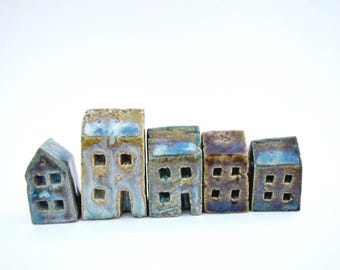 Puede incluir: Un conjunto de cinco pequeñas figuras de casas de cerámica dispuestas en fila. Cada casa presenta un acabado esmaltado y texturizado en tonos azules, marrones y beige, con ventanas y puertas cuadradas recortadas en la estructura.