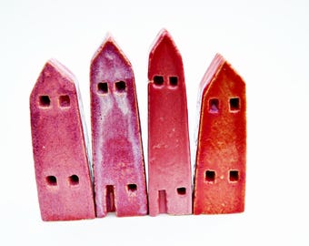 Puede incluir: Cuatro figuras de casas de cerámica altas y estrechas dispuestas en fila. Cada pieza presenta un techo puntiagudo, pequeñas ventanas cuadradas recortadas y una abertura de puerta rectangular. Las casas tienen tonos de rojo, magenta y rosa.
