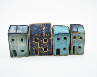 Puede incluir: Cuatro pequeñas figuras de casas de cerámica dispuestas en fila. Cada pieza presenta un acabado esmaltado en tonos azules, desde turquesa claro hasta azul marino profundo, con grietas visibles en el esmalte y aberturas cuadradas.