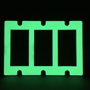 May include: A green glow-in-the-dark wall plate with three openings for light switches. The plate has a smooth, flat surface and is made of plastic. The plate is designed to be installed on a wall.