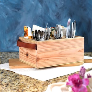 May include: A wooden cutlery organizer with a leather handle. The organizer has multiple compartments for storing forks, spoons, and knives. It is sitting on a countertop with a white napkin and a plate with a pink napkin and flowers.