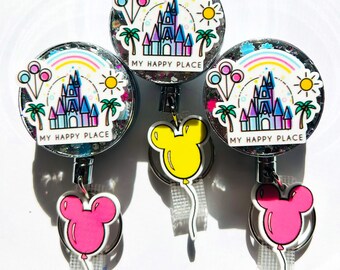 May include: Three retractable badge holders with a glittery, circular design. Each features a castle, rainbow, sun, and palm trees, with the text "MY HAPPY PLACE." Attached are Mickey Mouse balloon charms in pink and yellow.