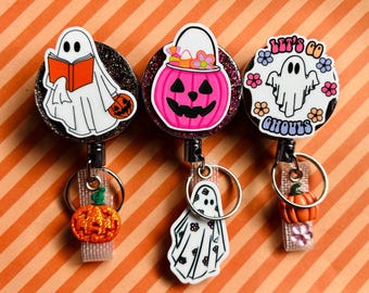 May include: Three circular badge reels with glittery black bases. The first features a ghost reading an orange book with a pumpkin charm. The second shows a pink jack-o'-lantern bucket with a ghost charm. The third displays a ghost with the text LET'S GO GHOULS and a pumpkin charm.