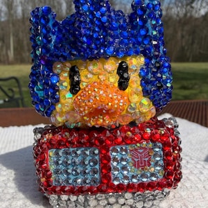 May include: A bedazzled figurine of a character with a blue head, yellow face, and red body. The figurine is covered in sparkling rhinestones. The character has black eyes and a red emblem on the chest.