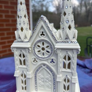 May include: A white ceramic church figurine with two spires, adorned with sparkling rhinestones. The church features arched windows, a decorative rose window, and a detailed front door. The figurine is placed on a purple surface.