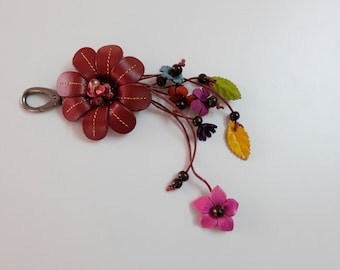 Könnte beinhalten: Ein Schlüsselanhänger aus burgunderfarbenem Leder in Form einer Blume mit einem silbernen Metallverschluss. Die Blume hat eine Perlenmitte und ist mit kleineren Blumen und Blättern in verschiedenen Farben verziert, darunter rosa, gelb, grün und blau.