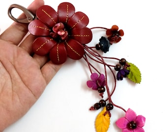 Könnte beinhalten: Ein Schlüsselanhänger aus burgunderfarbenem Leder in Form einer Blume mit einem Metallverschluss. Der Schlüsselanhänger hat eine Ansammlung kleinerer Blumen und Blätter in verschiedenen Farben, darunter rosa, lila, grün und gelb.