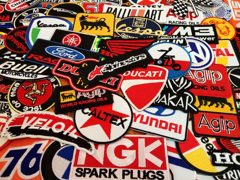 May include: A collection of colorful embroidered patches featuring various logos and brand names, including Vespa, Ford, Ducati, Agip, Caltex, and Hyundai. The patches are arranged in a random pattern and are perfect for adding a touch of personality to jackets, bags, or other items.