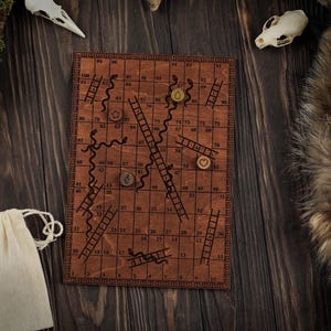Pode incluir: Um jogo de tabuleiro de Cobras e Escadas de madeira com detalhes gravados a laser. O tabuleiro apresenta números, cobras, escadas e bordas decorativas. Inclui peças de jogo de madeira e um saco com cordão. O jogo está sobre uma superfície de madeira escura.