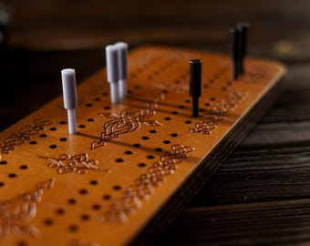 May include: A wooden cribbage board with decorative floral patterns and peg holes. The board is a warm brown color and has several pegs inserted, including white and black pegs. The pegs are cylindrical and stand upright.