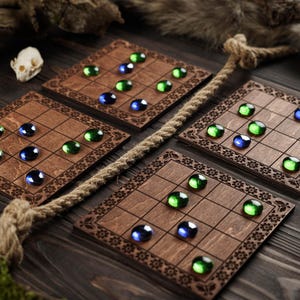 Puede incluir: Cuatro tableros de juego de madera con un patrón de cuadrícula, cada uno con canicas de vidrio verdes y azules. Los tableros tienen un borde floral decorativo y están dispuestos sobre una superficie de madera oscura.