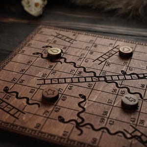 May include: A Snakes and Ladders board game made of wood. The board features a grid with numbers, ladders, and snakes. Wooden game pieces with heart, star, lightning bolt, and water drop symbols are on the board.