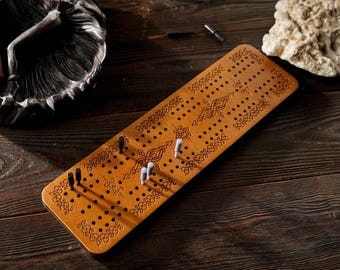 May include: A wooden cribbage board with decorative etched patterns and numerous holes for pegs. The board is light brown and has pegs in various colors. The image also shows an ashtray and a rock.
