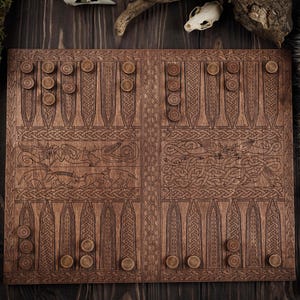 Puede incluir: Un tablero de backgammon de madera con intrincados diseños tallados y fichas de juego de madera. El tablero presenta ilustraciones detalladas de animales y patrones de nudos celtas. Las fichas son circulares y marrones. El tablero es de color marrón intenso.