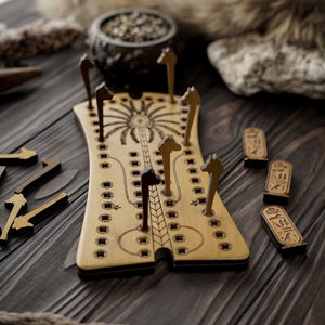 Può includere: Un tabellone da gioco in legno con un disegno centrale di una palma e un serpente. Il tabellone ha una serie di buchi e piste per i pezzi da gioco. Ci sono cinque pezzi da gioco in legno a forma di cavalli e tre tessere in legno con la parola "Osiris" scritta su di esse.