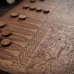 Puede incluir: Un tablero de backgammon de madera con intrincados diseños grabados y fichas de juego de madera. El tablero presenta ilustraciones detalladas, incluyendo patrones de nudos celtas y figuras. Las fichas son circulares, con una combinación de madera clara y oscura.