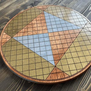 May include: A round wooden board game with a geometric design. The board features a grid pattern with copper, gold, and silver-colored sections. The board is approximately 18 inches in diameter.