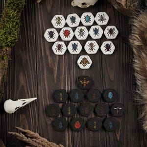 Puede incluir: Un conjunto de fichas de juego hexagonales con ilustraciones de insectos. Las fichas tienen dos esquemas de color: blanco con insectos negros y rojos, y negro con diseños de insectos dorados, azules y rojos. Dispuestas sobre una superficie de madera oscura.