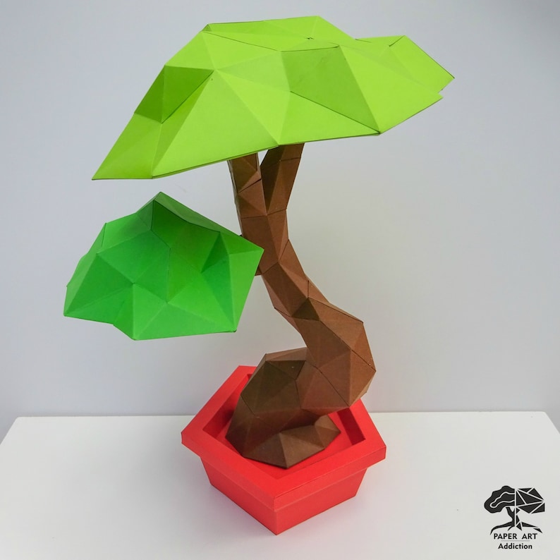 May include: A geometric paper bonsai tree sculpture. The tree has a brown trunk, two green leaf sections, and sits in a red square pot. The sculpture is made of folded paper and has a modern, angular design. The logo "PAPER ART Addiction" is in the bottom right corner.