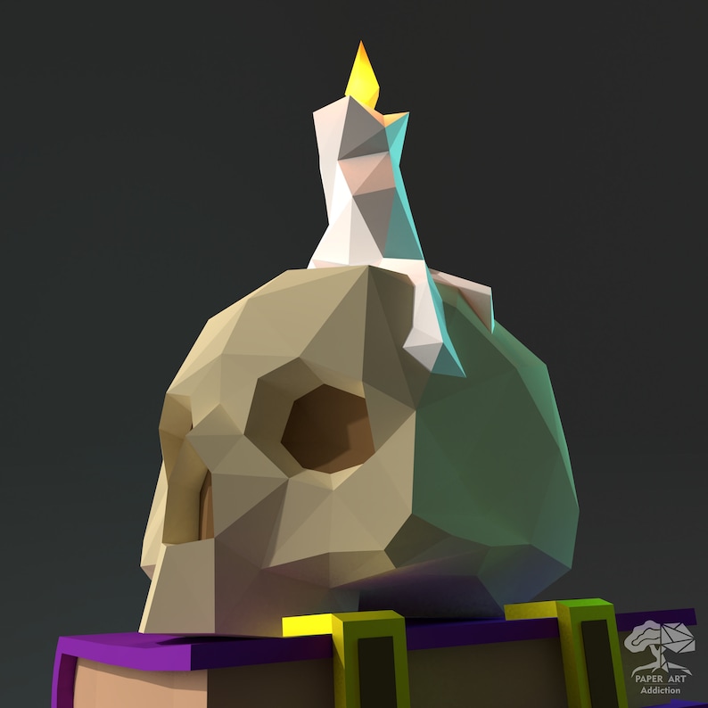 May include: A white candle with a yellow flame sits atop a beige papercraft skull. The skull is resting on a purple and yellow book with the words "PAPER ART Addiction" printed on the cover.