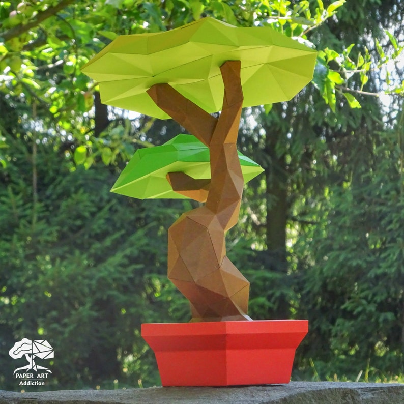 May include: A paper art bonsai tree with a geometric design. The tree has a brown trunk and light green leaves, set in a red pot. The background is a blurred view of green foliage. The logo "PAPER ART Addiction" is in the lower left corner.