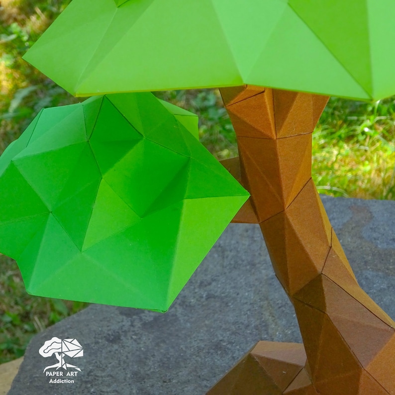 May include: A geometric paper art tree with a brown trunk and bright green leaves. The tree is made of folded paper and has a modern, angular design. The tree is sitting on a gray stone surface. The logo "Paper Art Addiction" is visible.