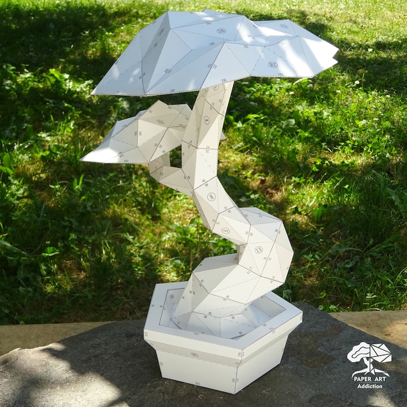 May include: A white paper bonsai tree sculpture with geometric shapes. The tree has a detailed trunk, branches, and a full canopy, all made from folded paper. The sculpture sits in a square pot, also made of white paper. The background is green grass.