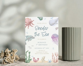 Puede incluir: Una tarjeta de invitación azul y blanca con el texto "Oneder the Sea" e ilustraciones de criaturas marinas, incluyendo un delfín, un pulpo, una medusa y una ballena. La invitación es para una fiesta de primer cumpleaños.