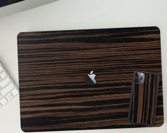 Puede incluir: Una funda para portátil con textura de madera y una funda para teléfono a juego. La funda para portátil cubre la parte superior de un MacBook, con un patrón de rayas marrón oscuro y negro. La funda del teléfono tiene un diseño similar.