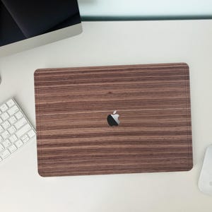 Könnte beinhalten: Eine braune Laptop-Hülle aus Holz mit einem schwarzen Apple-Logo in der Mitte. Die Hülle hat ein Holzmaserungsmuster und ist von einer weißen Tastatur, Maus und einem Computermonitor umgeben. Die Hülle ist rechteckig.