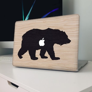 May include: A laptop case with a light wood grain finish. A black silhouette of a bear is centered on the case, with a white Apple logo incorporated into the bear's body. The laptop is on a white desk.