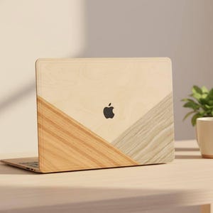 May include: A laptop with a wooden finish, featuring a black Apple logo. The design incorporates light and medium brown wood tones in a geometric pattern. A white mug, plant, and notebook are on the desk.