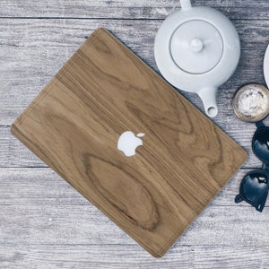Könnte beinhalten: Eine hellbraune Laptop-Hülle aus Holz mit weißem Apple-Logo. Die Hülle hat ein Holzmaserungsmuster. Eine weiße Teekanne, Sonnenbrille und eine kleine Schale sind ebenfalls sichtbar.