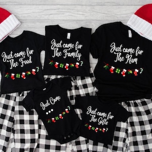 May include: A collection of black t-shirts and a baby bodysuit featuring white text and a colorful Christmas stocking and gift graphic. The shirts display phrases including Just came for The Food, The Family, The Rum, and The Gifts, with one bodysuit reading Just Came. Two red and white Santa hats are included.