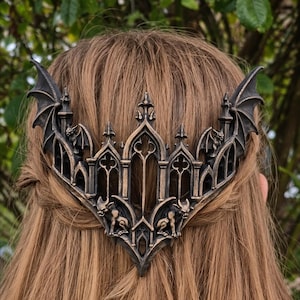 May include: A bronze-coloured hair accessory featuring a detailed gothic architectural design with pointed arches, spires, and small gargoyle figures. The piece includes wing-like extensions on both sides and has a dark, weathered finish.