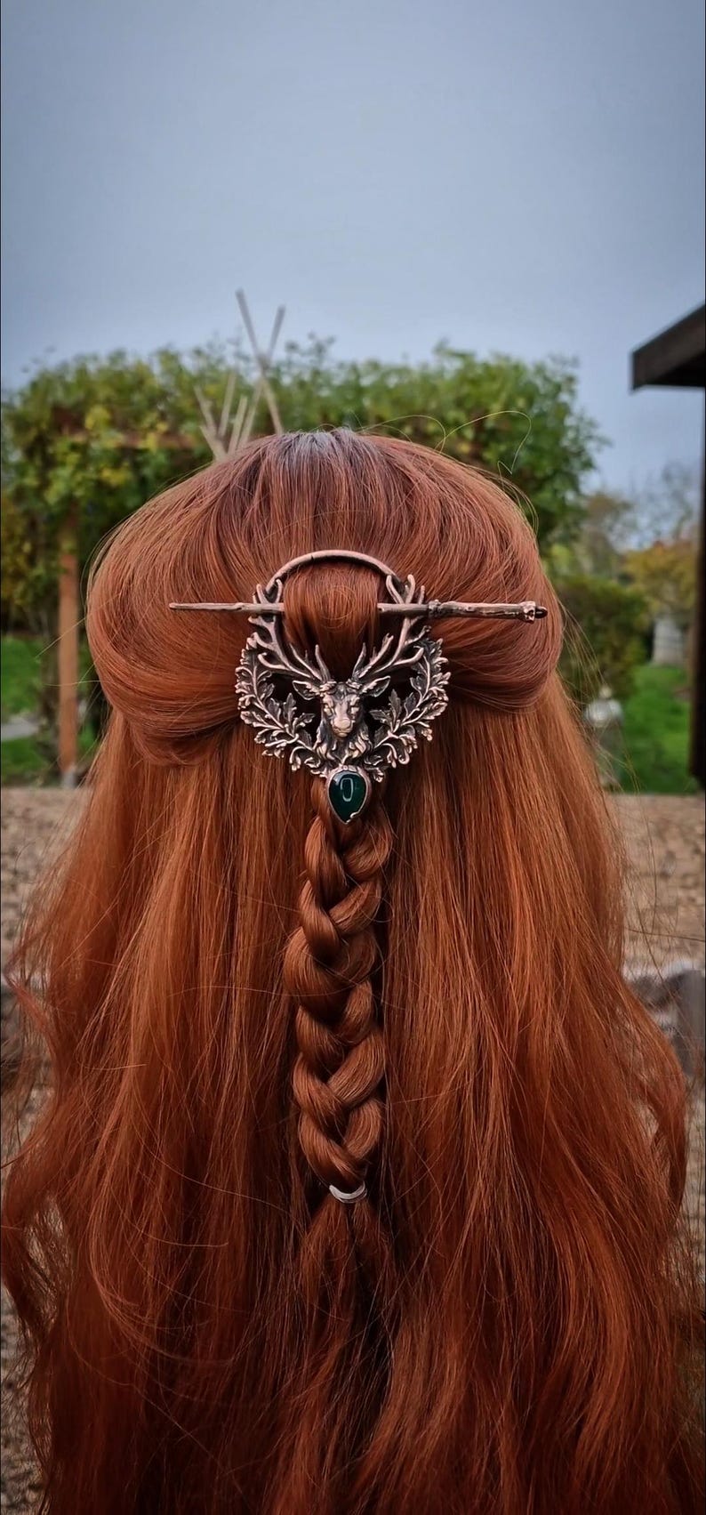 May include: A copper-toned hair accessory featuring a deer head design with a green gemstone. The hair piece is set against a backdrop of long, auburn hair styled in a braid.