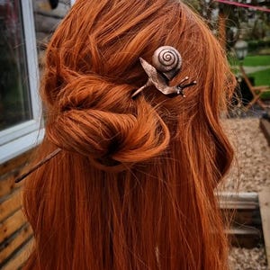 May include: A copper hair pin featuring a snail design. The snail is curled up in its shell and has a detailed texture. The pin is inserted into a woman's red hair.