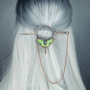 May include: A green and copper butterfly hair clip with a chain hanging from the bottom. The clip is attached to a woman's hair.