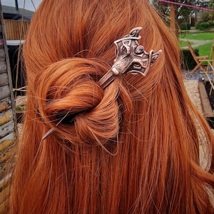 Könnte beinhalten: Eine Haarnadel aus Kupfer mit einem detaillierten Design eines Wolfskopfes mit einem grünen Edelsteinauge. Die Nadel befindet sich in den roten Haaren einer Frau.